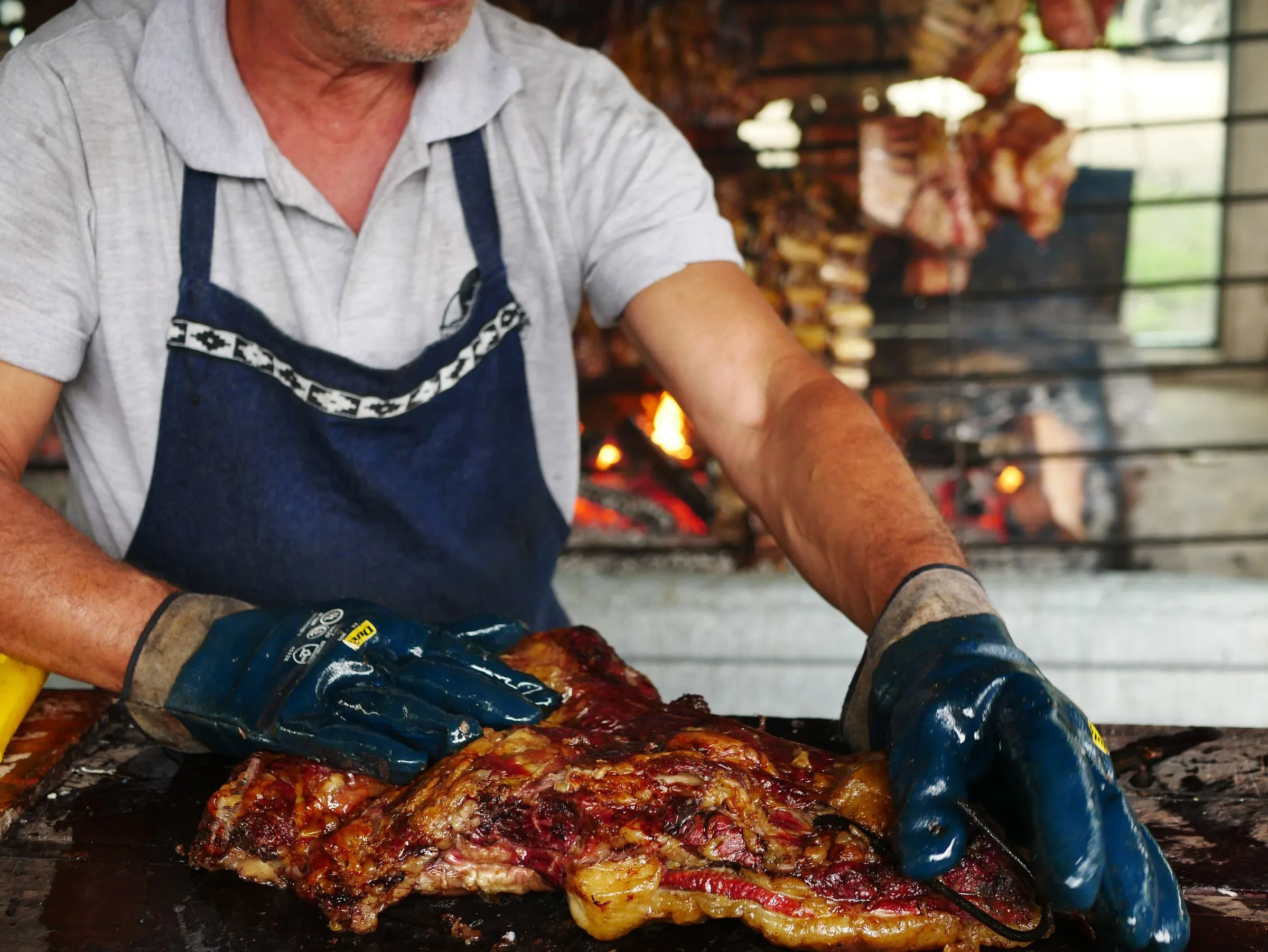 Manos de nuestro parrillero levantando una pieza de carne recién asada junto al fuego de la parrilla
